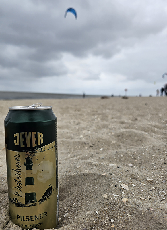 Eine Dose Jever Pilsener steht im Sand am Strand. Im Hintergrund sind Kitesurfer am bewölkten Himmel zu sehen. 