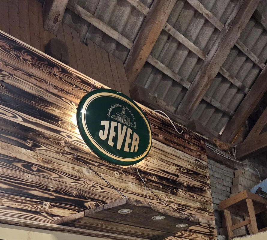  Ein beleuchtetes Jever-Schild hängt an einer Holzwand in einem rustikalen Raum mit Holzbalken und Ziegeldach.