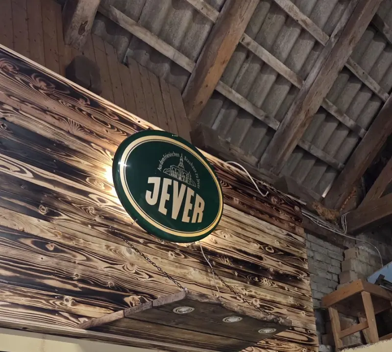 Ein beleuchtetes Jever-Schild hängt an einer Holzwand in einem rustikalen Raum mit Holzbalken und Ziegeldach.