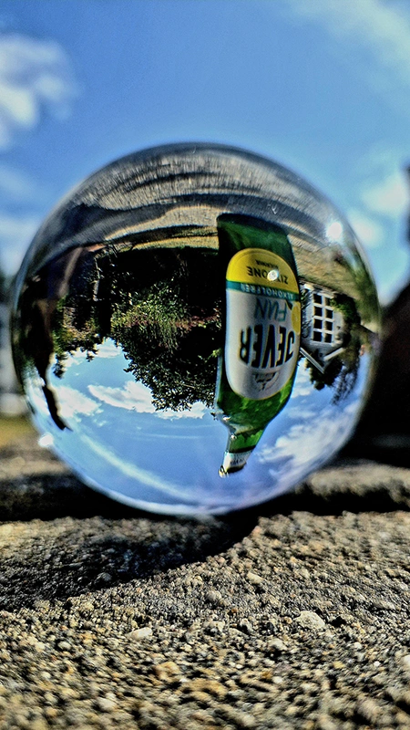 Eine Glaskugel reflektiert eine Flasche und eine Landschaft mit blauem Himmel. Die Szene wirkt surreal und verzerrt.