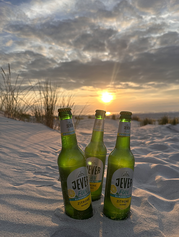 Drei Flaschen alkoholfreies Bier stehen im Sand, während die Sonne am Horizont untergeht. Wolken ziehen über den Himmel. 