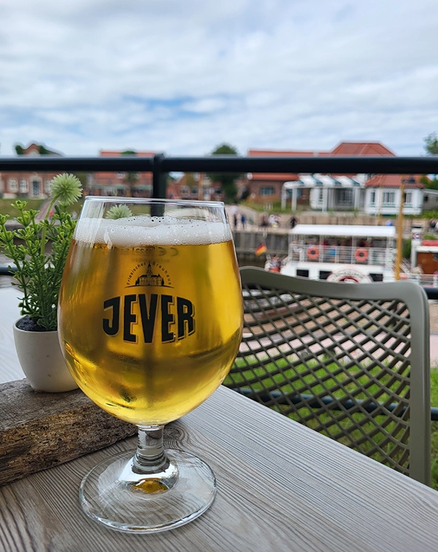 Ein Glas Jever-Bier auf einem Tisch, daneben eine kleine Pflanze. Im Hintergrund ein Hafen mit Booten und roten Ziegeldächern.