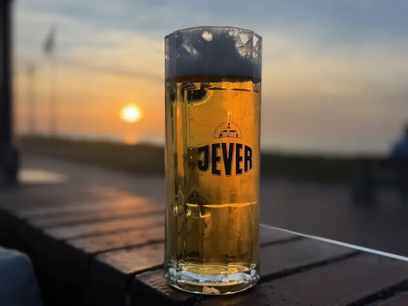 Ein Glas Bier mit Schaumkrone steht auf einem Tisch im Freien. Im Hintergrund geht die Sonne unter und taucht die Szene in warmes Licht.