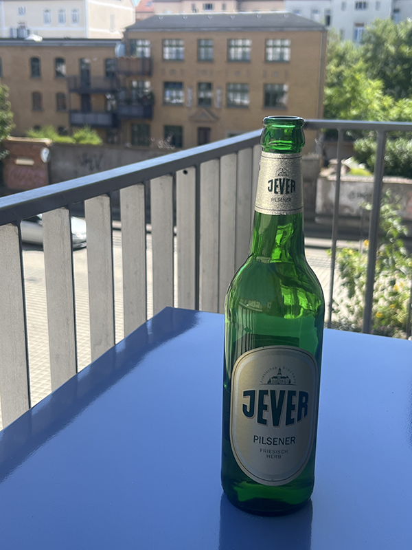 Eine grüne Bierflasche steht auf einem blauen Tisch auf einem Balkon. Im Hintergrund sind Gebäude und Bäume zu sehen.
