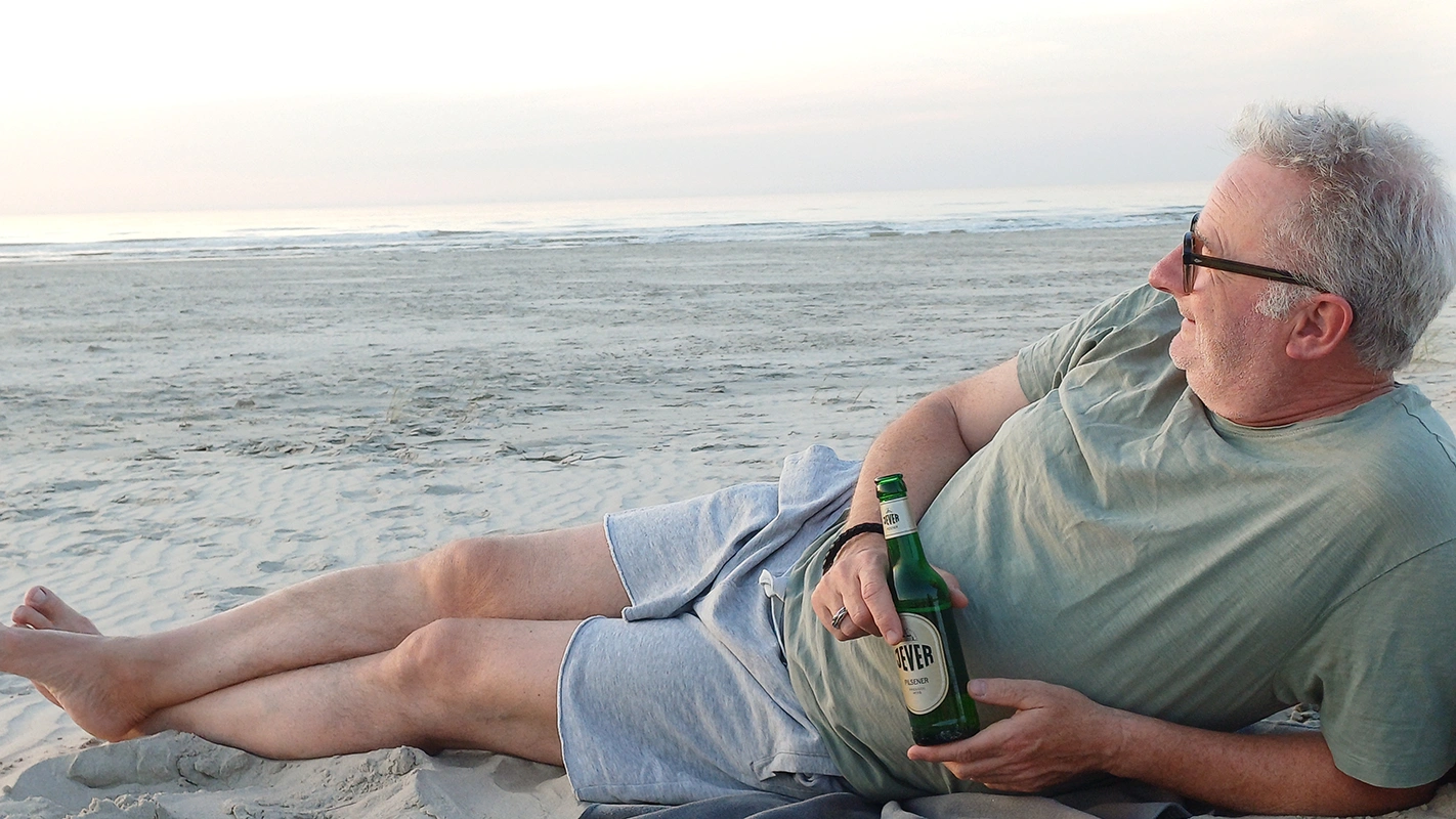 Ein Mann liegt entspannt am Strand, hält eine Bierflasche und schaut aufs Meer. Der Himmel ist leicht bewölkt. 