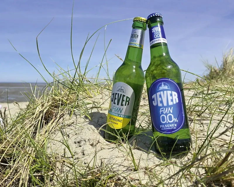 Zwei Flaschen alkoholfreies Bier stehen im Sand am Strand, umgeben von Gras, mit Blick auf das Meer unter klarem Himmel.
