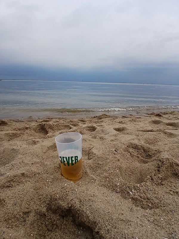 Ein Plastikbecher mit Getränk steht im Sand am Strand. Im Hintergrund das Meer unter einem bewölkten Himmel.