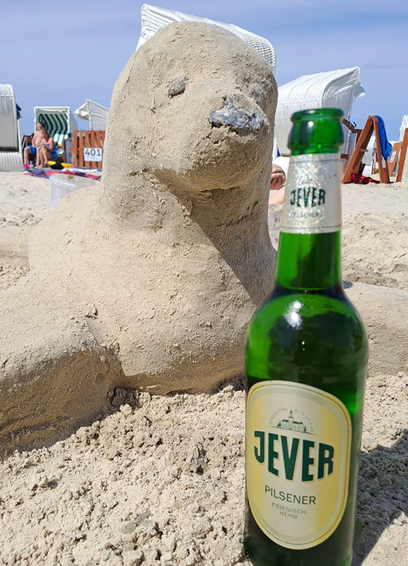 Eine Sandfigur in Form einer Robbe liegt am Strand. Daneben steht eine grüne Flasche Jever Pilsener auf dem Sand.