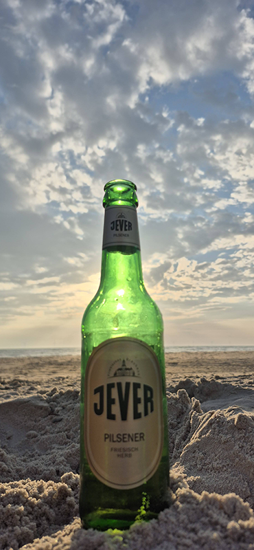 Eine grüne Jever Bierflasche steht im Sand vor einem bewölkten Himmel am Strand. Die Sonne scheint durch die Wolken. 
