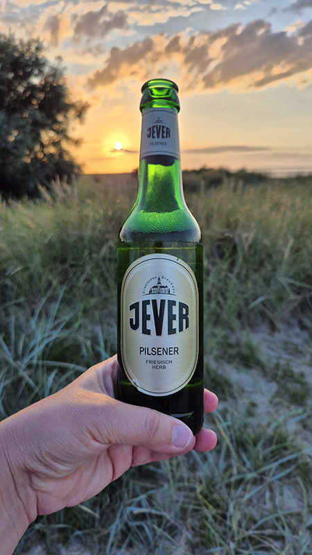 Eine Hand hält eine Flasche Jever Pilsener vor einem Sonnenuntergang. Im Hintergrund sind Dünen und ein bewölkter Himmel zu sehen.