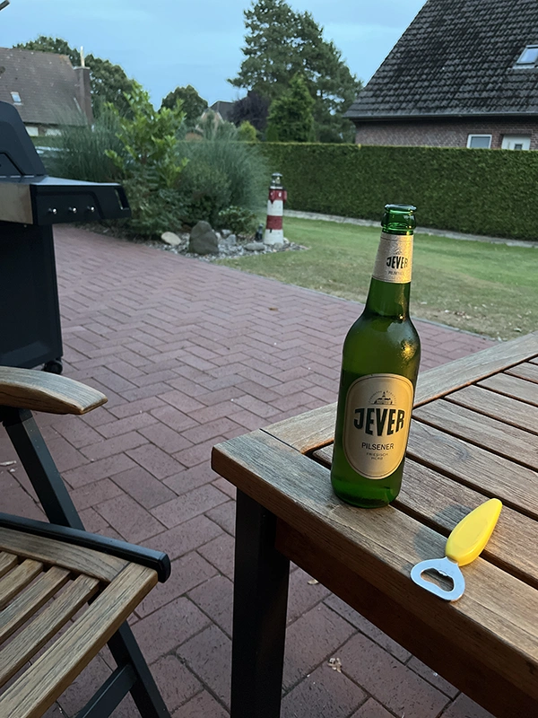 Im Vordergrund ist ein Holztisch mit einer Jever Bierflasche und Öffner. Im Hintergrund ist ein Grill und ein gepflegter Garten mit Hecke zu sehen.