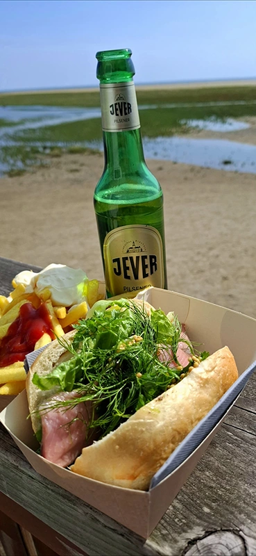 Eine Flasche Jever Pilsener steht neben einem Fischbrötchen und Pommes mit Ketchup und Mayo auf einem Holztisch am Strand.