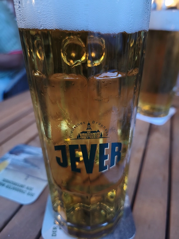 Ein Glas Jever Bier steht auf einem Holztisch, umgeben von Bierdeckeln. Die goldene Farbe des Bieres leuchtet im Abendlicht.