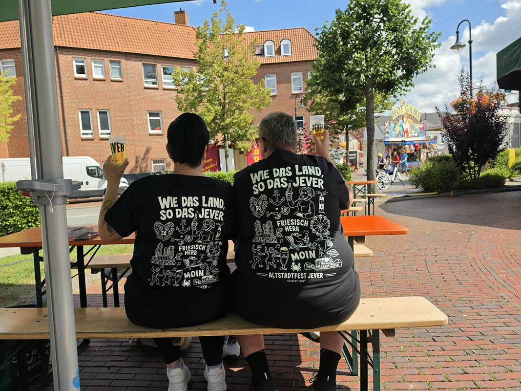 Zwei Personen sitzen auf einer Bank, tragen schwarze T-Shirts mit Aufdruck auf dem Rücken und halten Jever-Becher. Im Hintergrund ist ein Fest. 