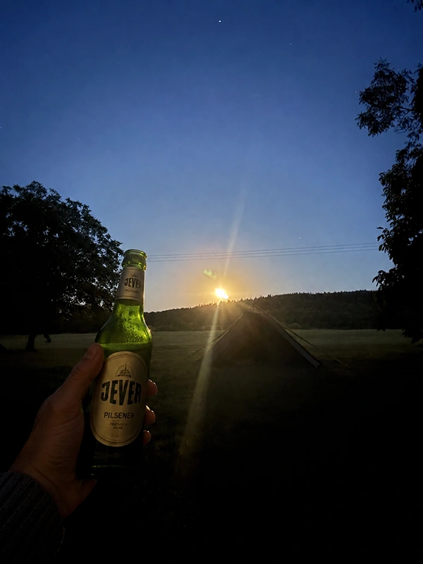 Eine Hand hält eine Flasche Jever Pilsener vor einem Sonnenuntergang auf einem Feld mit Bäumen und einem Zelt im Hintergrund. 