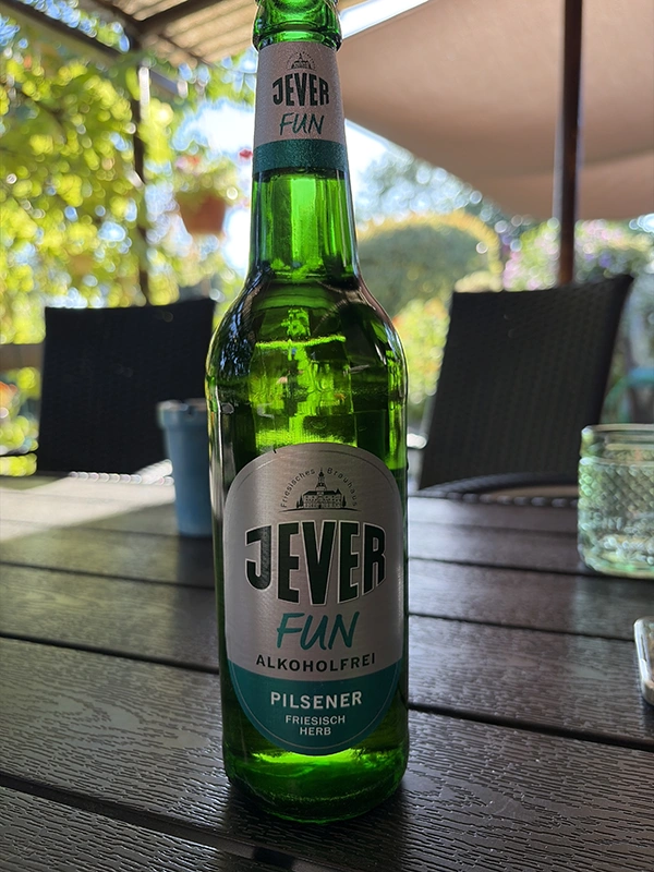 Eine grüne Flasche Jever Fun alkoholfreies Pilsener steht auf einem Holztisch im Freien. Im Hintergrund sind unscharfe Gartenmöbel.