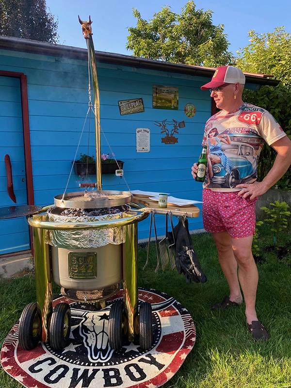Ein Mann steht neben einem Grill im Garten, trägt bunte Kleidung und hält eine Flasche Jever. Der Grill hat ein auffälliges Design. 