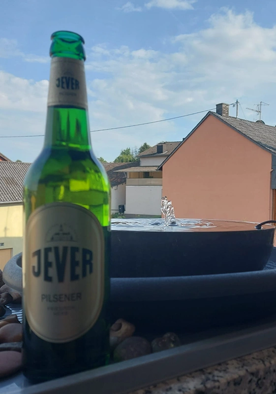 Ein Jever Pilsener steht auf einem Tisch mit Steinen. Dahinter sprudelt ein kleiner Brunnen, umgeben von Häusern.