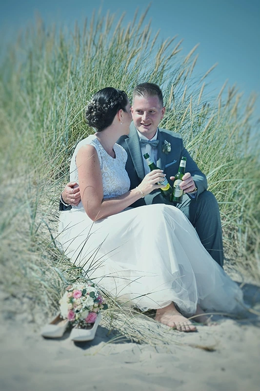 Ein Brautpaar sitzt am Strand im Sand, umgeben von Dünen. Beide halten eine Flasche Jever und lächeln einander an. 