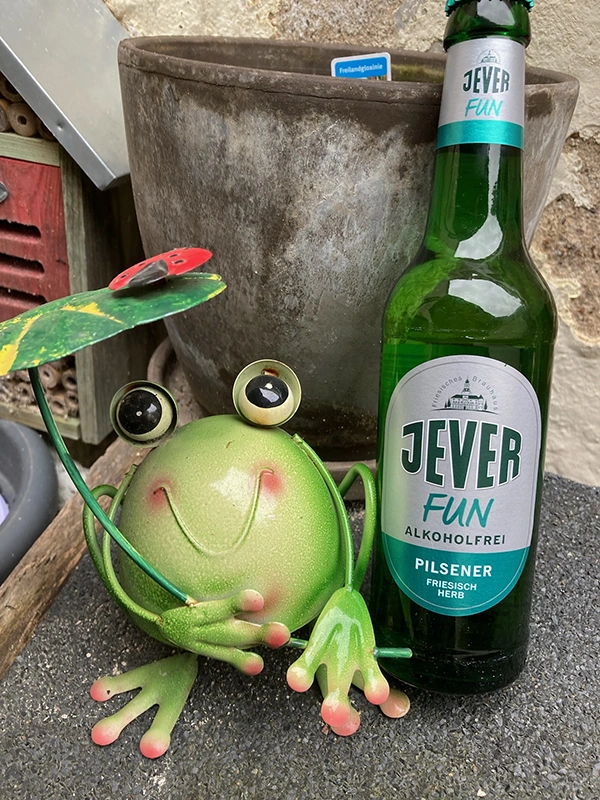 Ein grüner Frosch aus Metall hält ein Blatt mit einem Marienkäfer. Daneben steht eine Flasche Jever Fun alkoholfrei. 