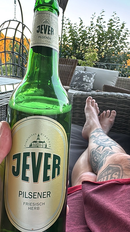 Nahaufnahme einer Jever Pilsener Flasche, gehalten von einer Person, die auf einer Terrasse mit Blick auf den Garten sitzt.