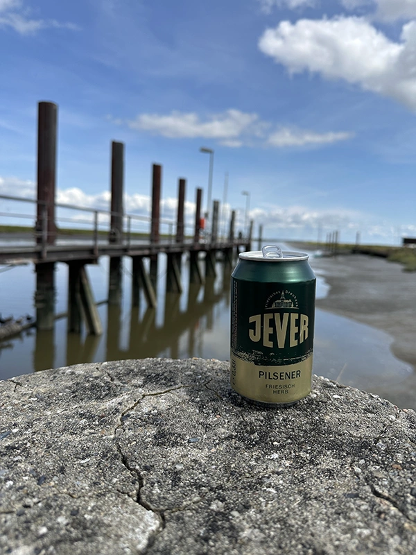 Eine Dose Jever Pilsener steht auf einem Betonpfeiler vor einem hölzernen Steg, der ins Wasser führt. Blauer Himmel im Hintergrund. 