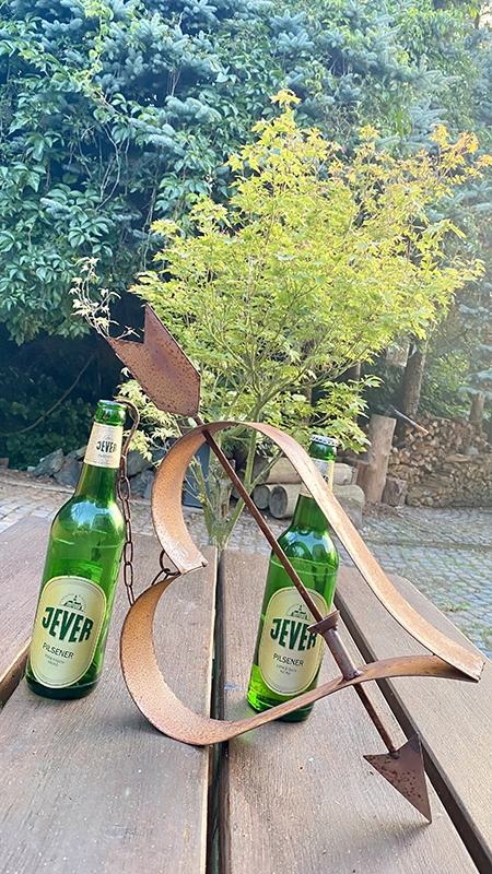 Zwei Jever Pilsener Flaschen stehen auf einem Holztisch. Daneben steht ein dekorativer Metallpfeil, der in einer Herzform steckt.