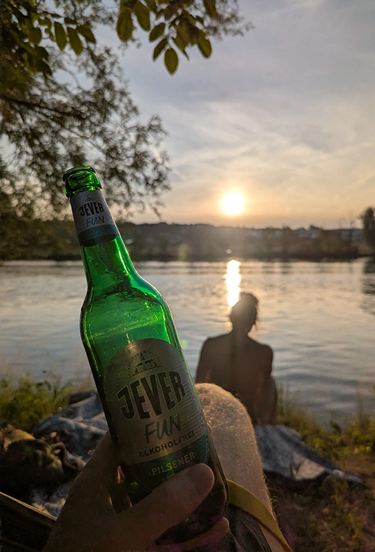 Eine Person hält eine Flasche Jefer Fun alkoholfreie am Flussufer, während die Sonne über dem Wasser untergeht. 