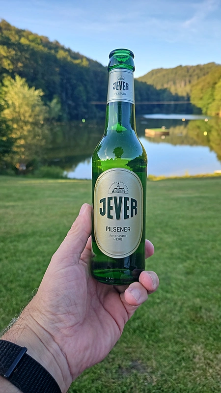 Eine Hand hält eine Flasche Jever Pilsener vor einer malerischen Seelandschaft mit bewaldeten Hügeln im Hintergrund. 