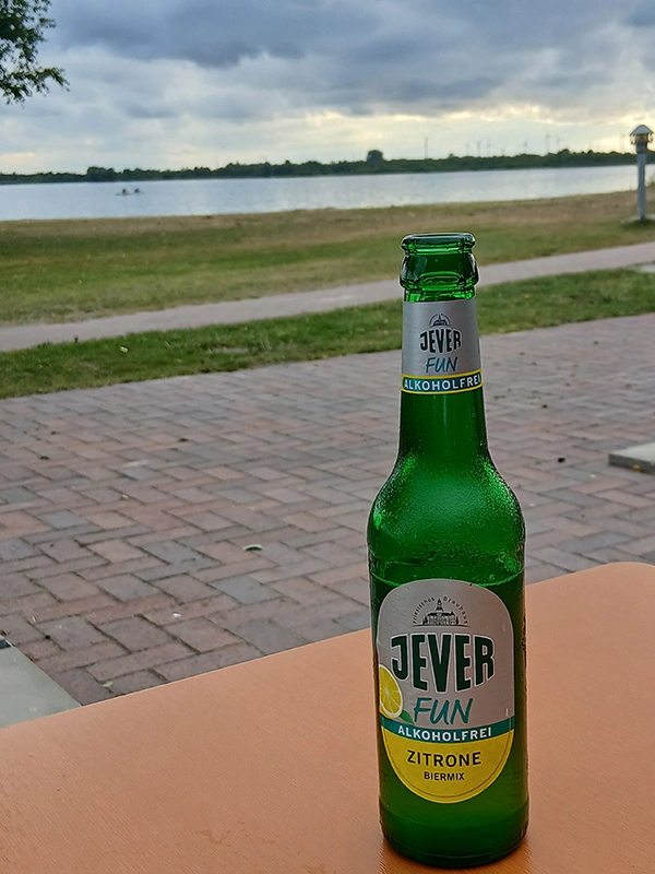Eine Flasche Jever Fun alkoholfrei mit Zitronengeschmack steht auf einem Tisch vor einem See mit bewölktem Himmel. 
