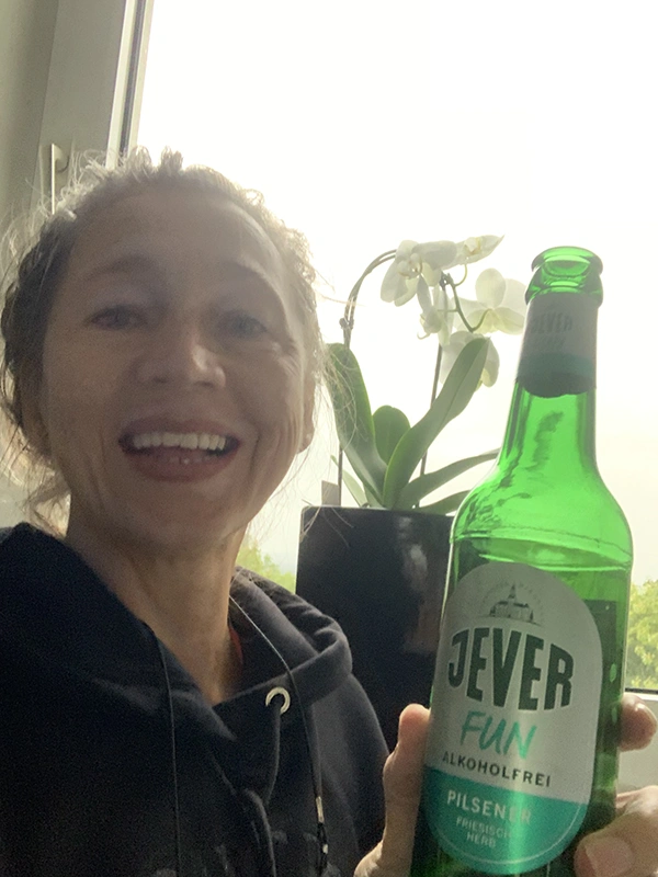 Frau lächelt in die Kamera und hält eine Flasche Jever Fun alkoholfreies Bier. Im Hintergrund steht eine weiße Orchidee.
