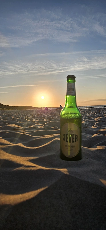 Eine Flasche Jever Pilsener steht im Sand am Strand, während die Sonne am Horizont untergeht und den Himmel in warmen Farben erleuchtet. 