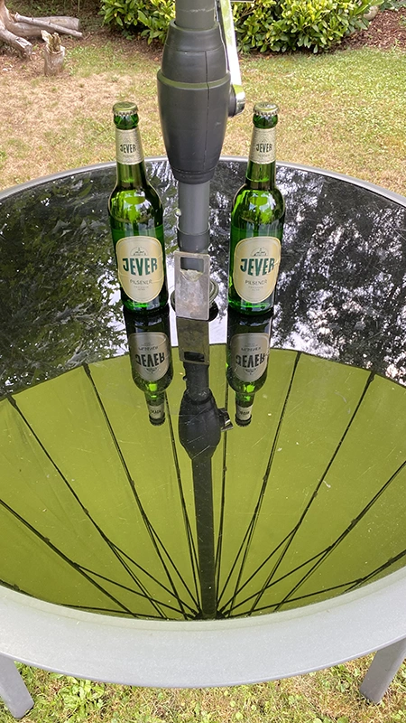 Zwei grüne Bierflaschen stehen auf einem runden Glastisch im Garten. Die Flaschen spiegeln sich auf der glänzenden Oberfläche.