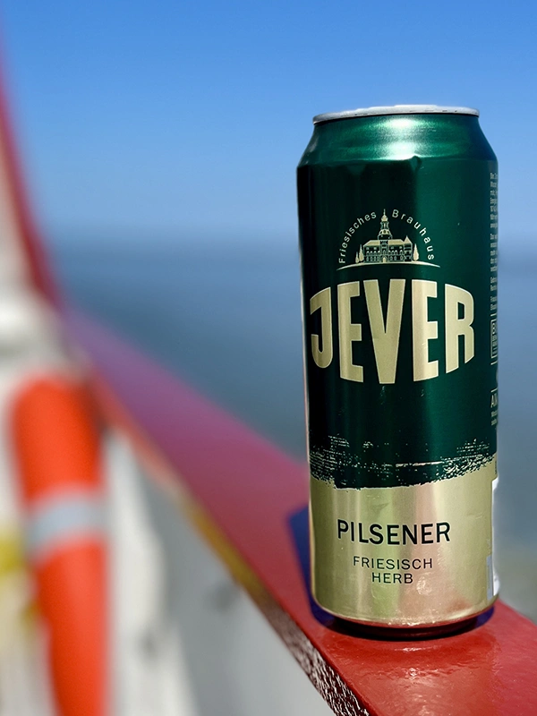 Eine grüne Dose Jever Pilsener steht auf einem roten Geländer, im Hintergrund ist verschwommen das Meer und der blaue Himmel zu sehen.