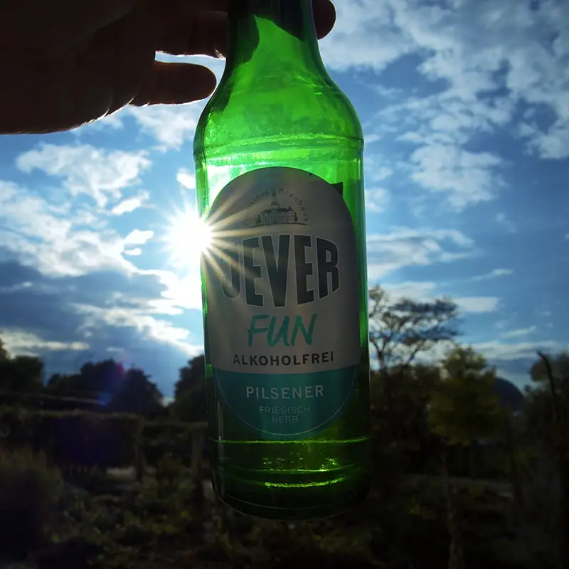 Eine Flasche Jever Fun alkoholfrei wird gegen den Himmel gehalten, die Sonne scheint durch das Glas. 