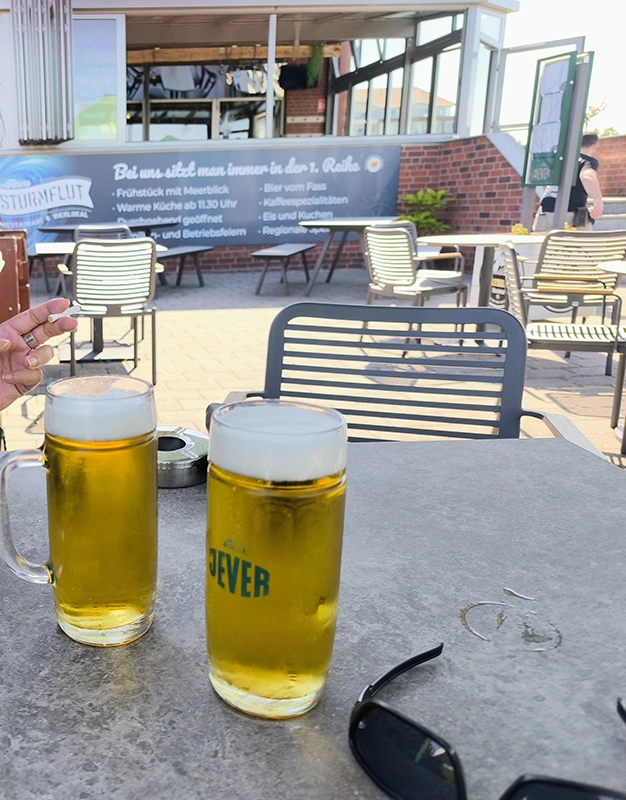 Zwei Gläser Jever Bier auf einem Tisch auf einer Café Terrasse, daneben liegt eine Sonnenbrille. 