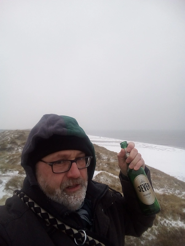 Mann mit Brille hält eine Jever Flasche in einer verschneiten Dünenlandschaft am Meer.