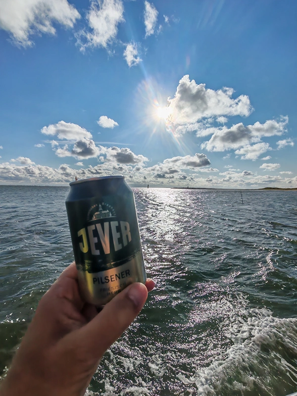 Eine Hand hält eine Jever Dose vor dem Meer. Sonne und Wolken spiegeln sich auf dem Wasser.