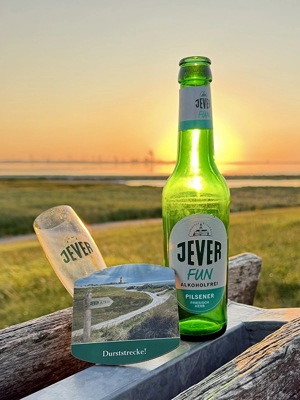 Eine Flasche Jever Fun steht mit Jever Glas und Bierdeckel auf einer Bank. Im Hintergrund liegt die Küste im Sonnenuntergang.