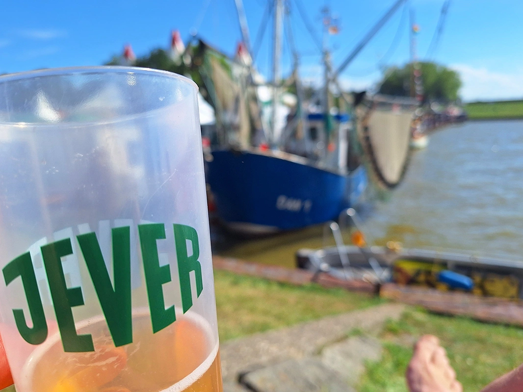Ein gefüllter Bierbecher von Jever wird vor einem Fischkutter am Wasser gehalten. Im Hintergrund liegen Hafen und Deich.