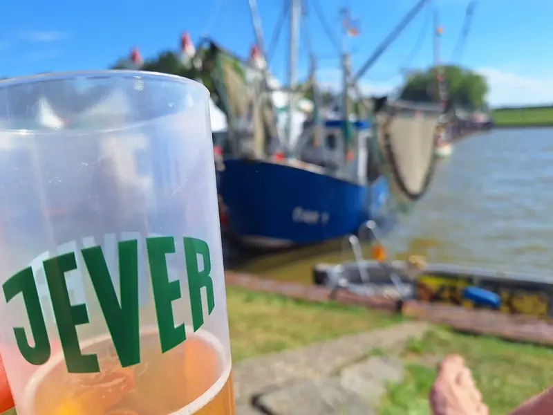 Ein gefüllter Bierbecher von Jever wird vor einem Fischkutter am Wasser gehalten. Im Hintergrund liegen Hafen und Deich.