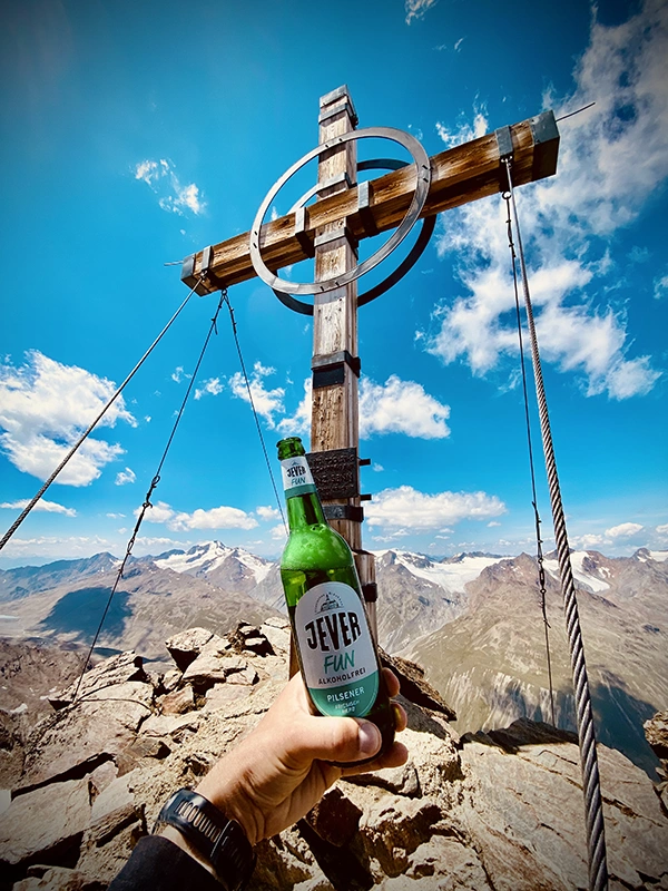 Eine Hand hält eine Flasche Jever Fun vor einem Gipfelkreuz. Im Hintergrund erstreckt sich eine Berglandschaft.