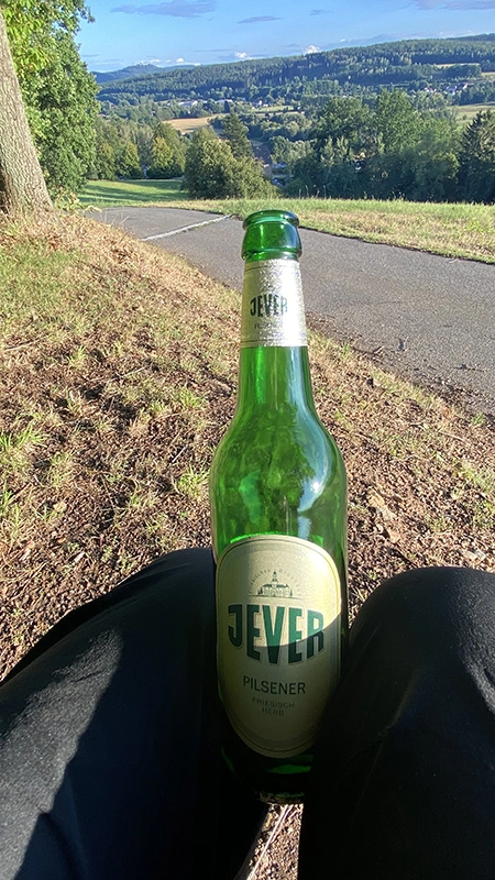 Eine Jever Flasche klemmt zwischen den Beinen einer sitzenden Person. Im Hintergrund liegt eine grüne Hügellandschaft.