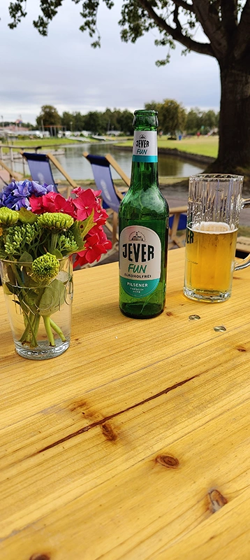 Jever Fun Flasche und Bierglas stehen neben Blumen auf einem Holztisch. Im Hintergrund liegt ein Gewässer.