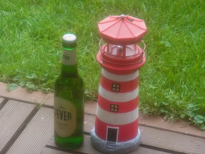 Eine Jever Flasche steht neben einem rot weißen Leuchtturmmodell auf einer Holzterrasse.