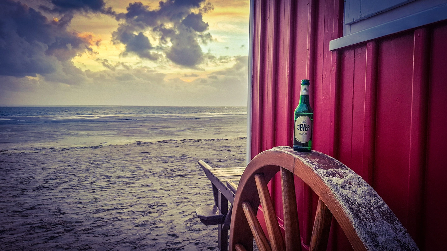 Eine Jever Flasche steht auf einem Holzrad vor einer roten Hütte. Im Hintergrund liegen Strand und Meer.