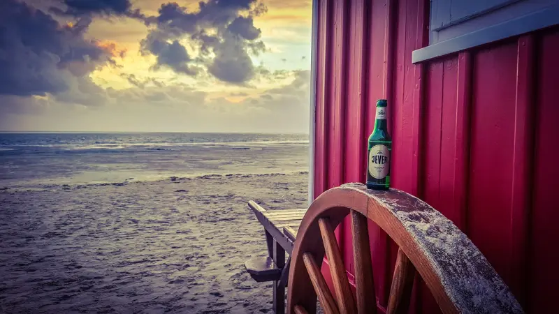 Eine Jever Flasche steht auf einem Holzrad vor einer roten Hütte. Im Hintergrund liegen Strand und Meer.