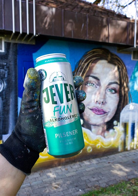 Eine Hand mit Arbeitshandschuh hält eine Dose Jever Fun. Im Hintergrund befindet sich ein gemaltes Graffiti Frauenporträt.