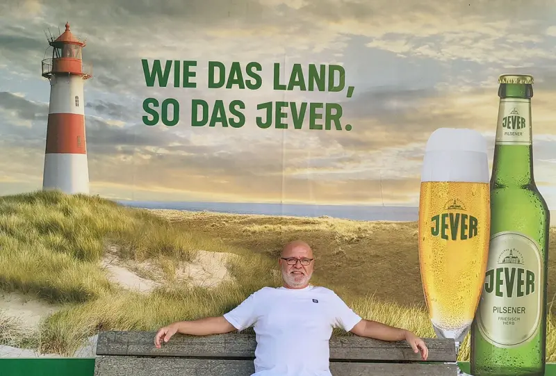 Ein Mann sitzt vor einer Jever Werbewand mit Leuchtturm, Bierglas und Flasche in einer Dünenlandschaft.