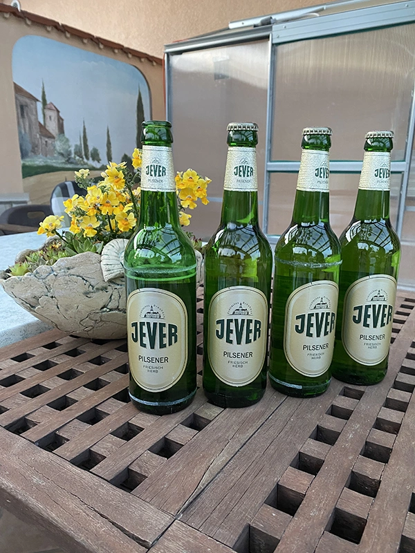 Vier Jever Flaschen stehen auf einem Holztisch neben gelben Blumen. Im Hintergrund befindet sich eine Terrasse.